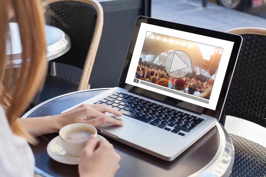 Person sitting outside at a small black round table with a laptop and drinking coffee while watching a video on a website