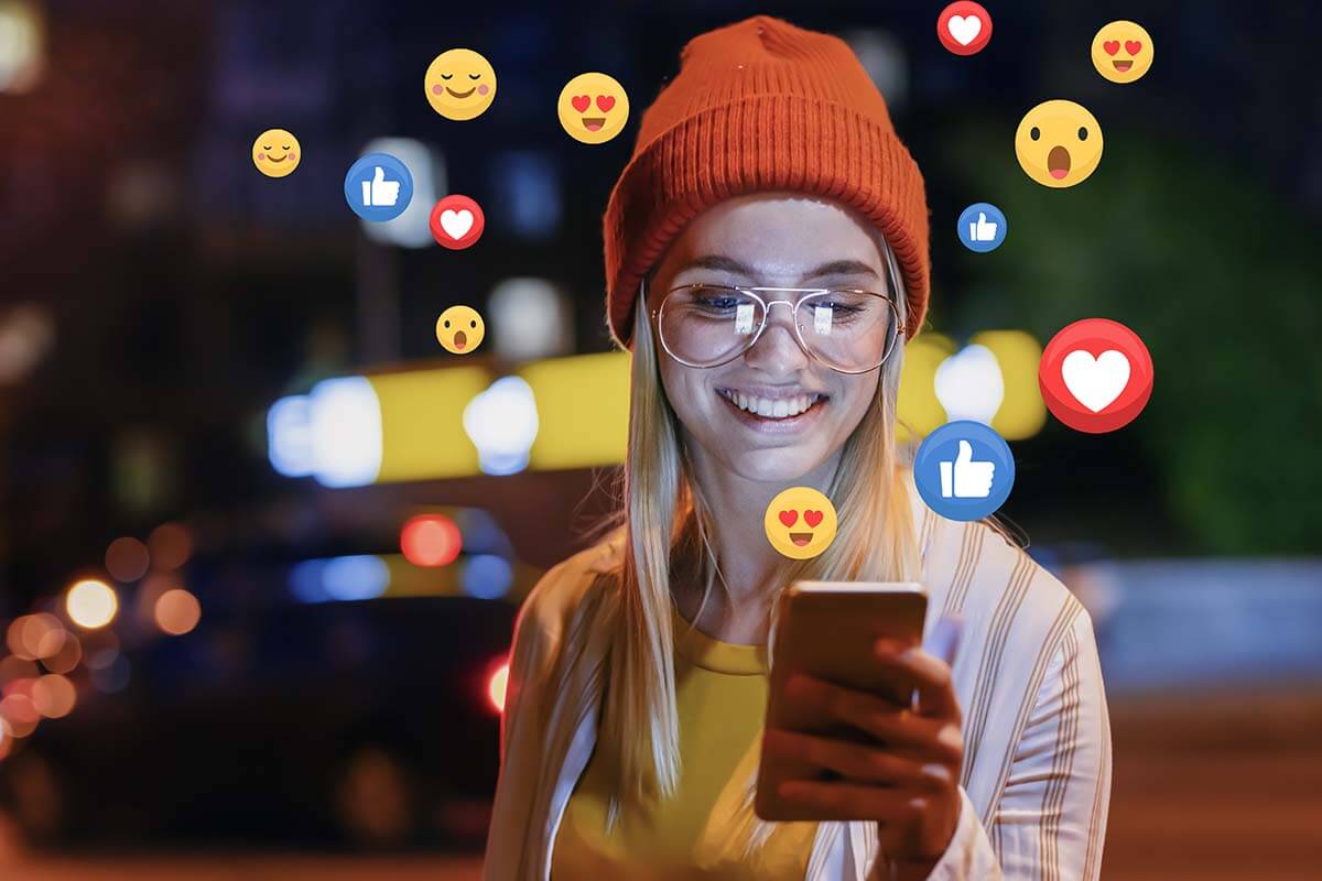 A beautiful young woman blogger, vlogger or influencer is receiving emoji and emoticon reactions in her mobile smart phone device while making a post, sharing or video logging on social media