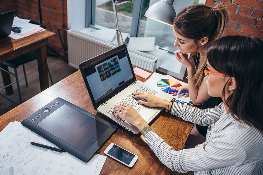 Two digital marketing professionals sitting at a wooden desk with a laptop and smart devices creating a unique website design for a client in Champaign, IL.