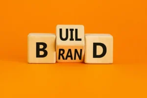 Right Click Digital, Inc. – Wooden letter blocks arranged on an orange background spell “BUILD” above “BRAND,” symbolizing the concept of building a strong brand identity.
