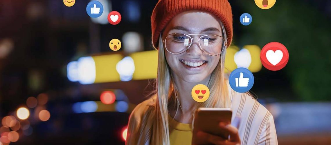 A beautiful young woman blogger, vlogger or influencer is receiving emoji and emoticon reactions in her mobile smart phone device while making a post, sharing or video logging on social media